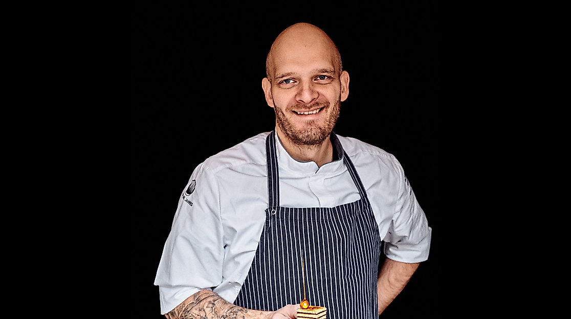 Björn Juhnke präsentiert festliches Weihnachtsdessert im Kochoutfit vor dunklem Hintergrund