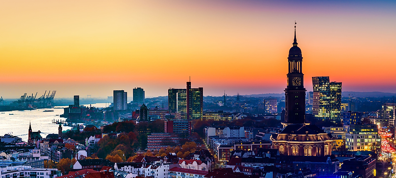 Panorama von Hamburg bei Sonnenuntergang mit beleuchtetem Michel-Turm und Hafen im Hintergrund.