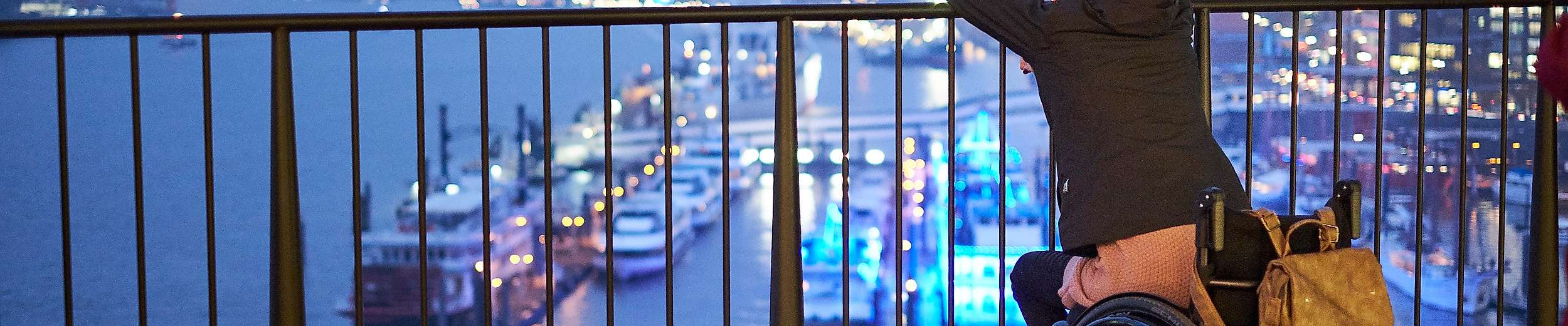 Rollstuhlnutzerin blickt abends von barrierefreier Plattform auf die beleuchtete Elbphilharmonie und den Hafen.