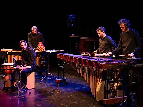 Three musicians play various percussion instruments. They are all dressed in black and bathed in atmospheric stage lighting.