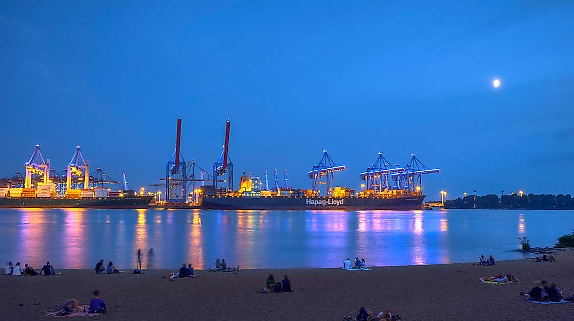 Abendstimmung am Elbstrand: Menschen sitzen entspannt am Sandufer, blicken auf den festlich beleuchteten Hamburger Hafen mit seinen Kränen und Lichtern – der Himmel ist tiefblau, der Mond steht über der Elbe.