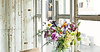 Vase mit frischen, bunt gemischten Blumen auf Holztisch vor Fensterfront in hellem Kreativraum