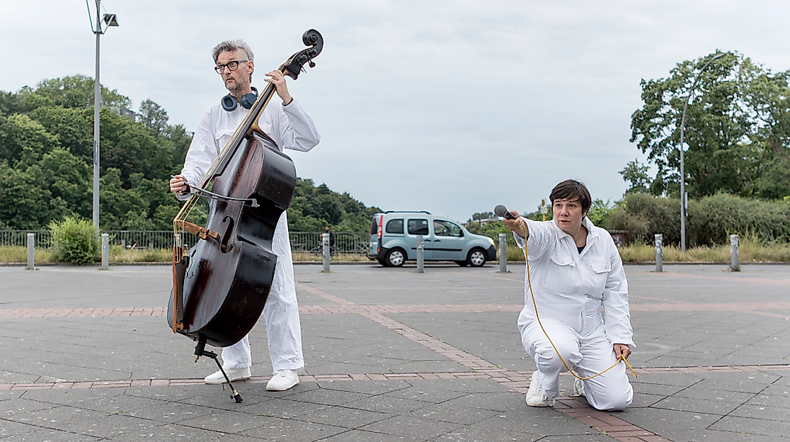In a parking lot, there are two people wearing white full-body suits and shoes. One person is playing the double bass, while the other is kneeling and holding out a microphone with one hand.