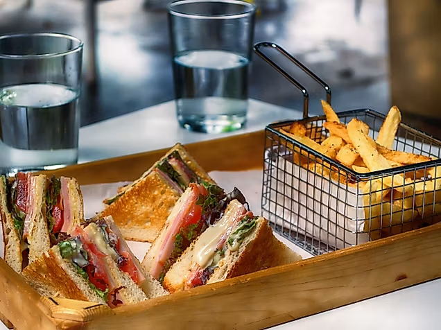Toast-Sandwiches mit Pommes in Metallkörbchen auf Holztablett, dazu zwei Gläser Wasser.