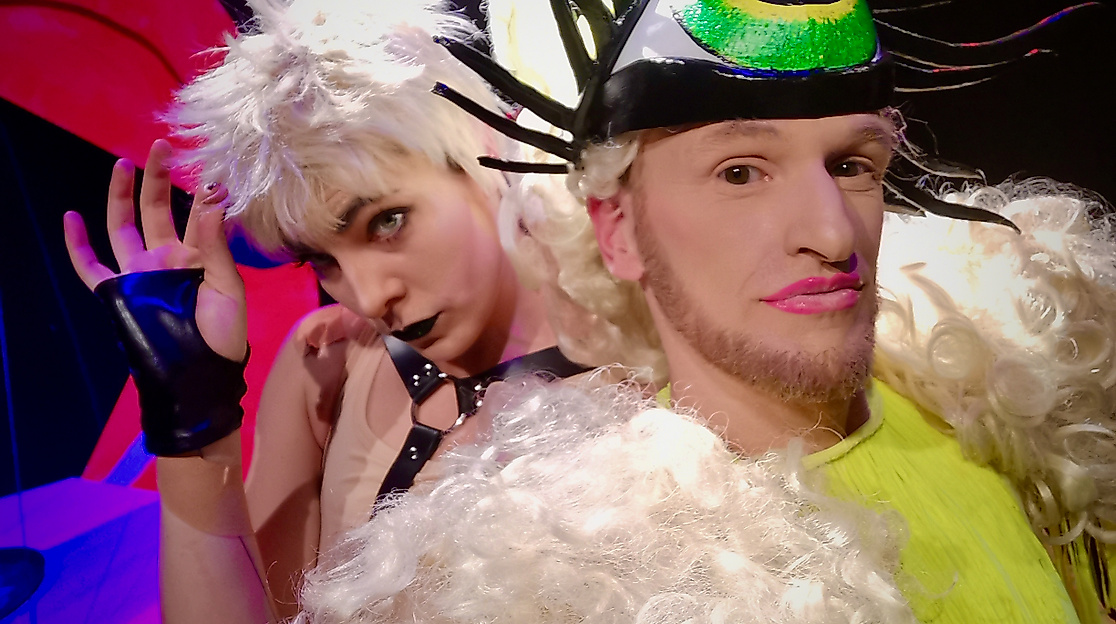 Zwei Performer*innen posieren in Kostümen. Eine Person trägt ein grünes Kostüm mit heller Locken-Perücke, dazu ein Stirnband mit einem großen Auge. Die Person dahinter trägt ein Harnessoutfit, ist jedoch nur halb zu sehen.