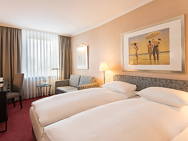 Helles Hotelzimmer mit Doppelbett, Sitzbereich und großem Fenster mit Vorhang.