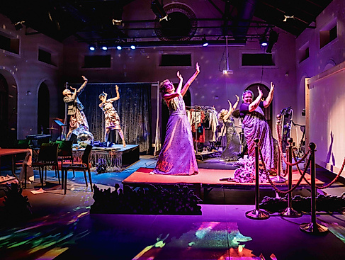 The performers dance around the stage to Bollywood music, making big gestures. There are colorful lights and the glow of a disco ball.