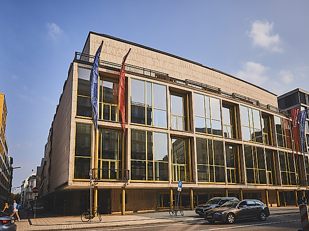 Moderne Glasfassade der Hamburgischen Staatsoper bei Tag mit Fahnen und Autos im Vordergrund.