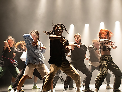 An energetic dance group performs a powerful choreography on stage under dramatic lighting.