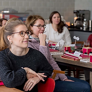 Teilnehmende hören aufmerksam im Lettering-Workshop zu, vor ihnen Tische mit Materialboxen und Getränken