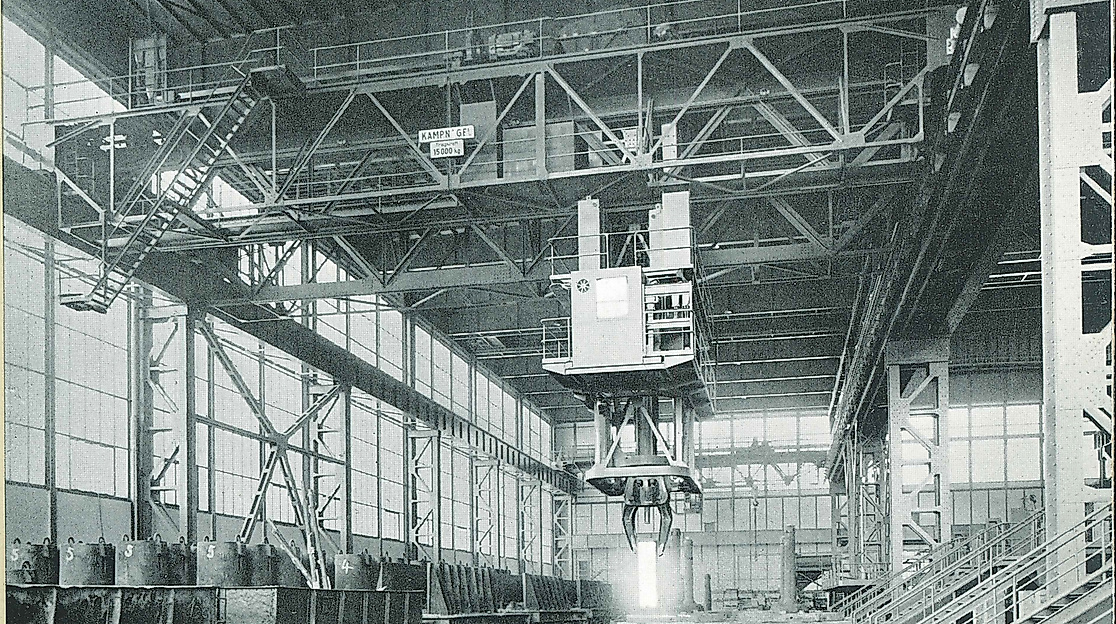 An old black and white photo of the former factory buildings of Kampnagel with scaffolding, a crane and metal barrels lined up.
