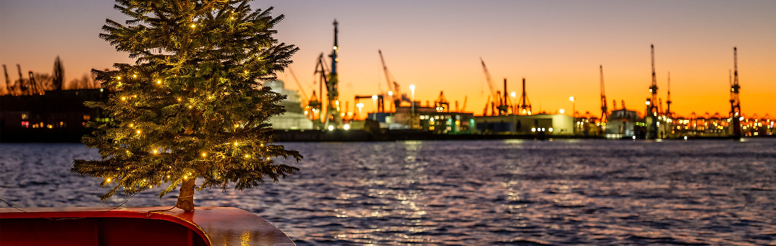Weihnachtstanne mit Lichterkette vor Sonnenuntergang an Bord eines Schiffes im Hamburger Hafen zur Adventszeit