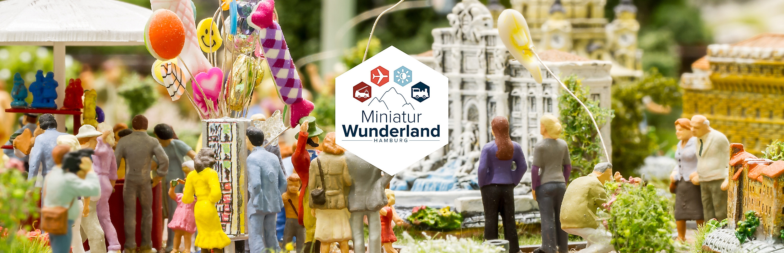 Bunte Miniaturszene mit Menschen, Luftballons und Modellgebäuden im Miniatur Wunderland Hamburg mit Logo des Miniatur Wunderlands Hamburg mit Symbolen für Bahn, Flugzeug, Sonne, Schiff und Lkw