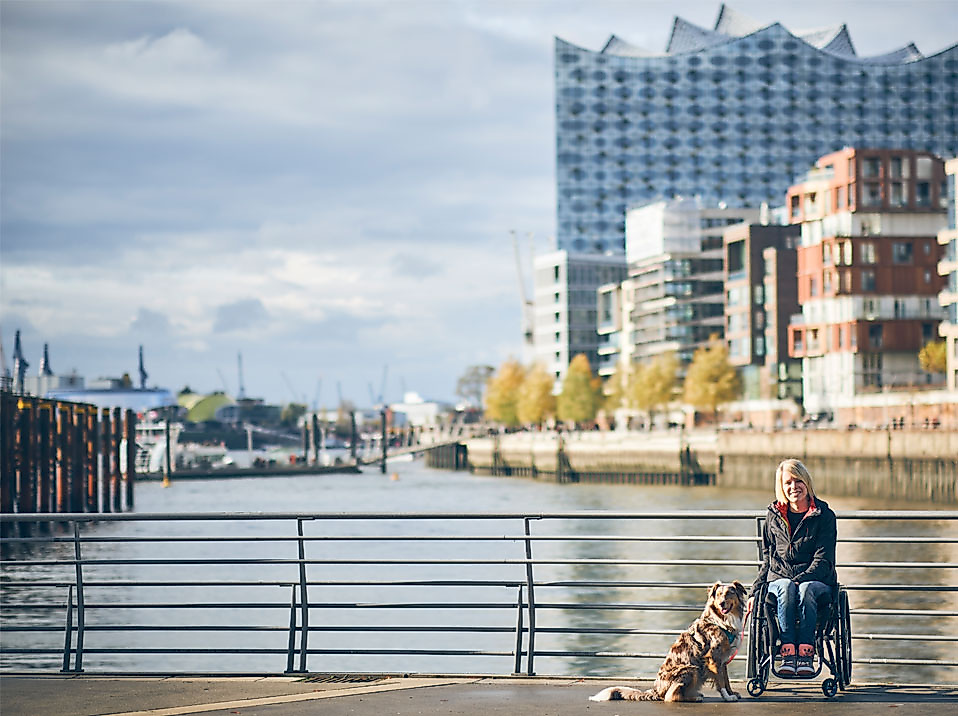 Rollstuhlfahrerin mit Assistenzhund in der HafenCity Hamburg, im Hintergrund Elbphilharmonie