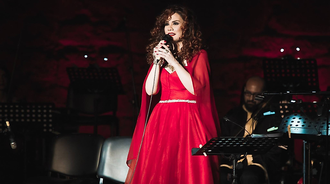 Lena Chamamyan, a woman with red curls, stands in a concert hall wearing a red dress and holding a microphone.