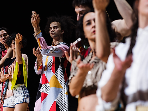 A group of dancers stand in a row and synchronously perform an expressive hand movement, focusing on one person in a colourful, geometrically patterned outfit.