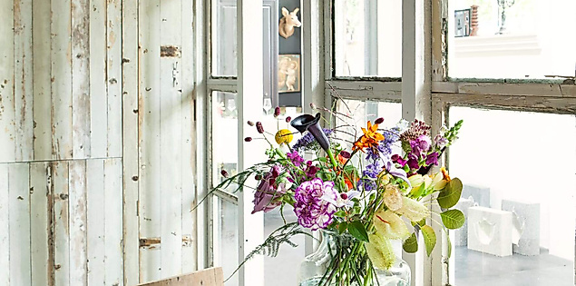 Vase mit frischen, bunt gemischten Blumen auf Holztisch vor Fensterfront in hellem Kreativraum