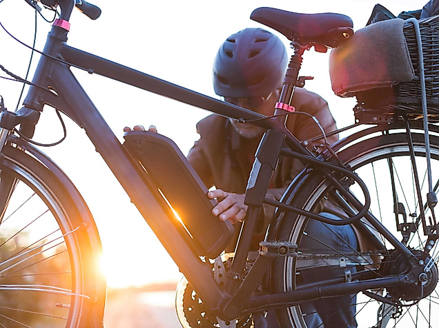 Person mit Helm prüft Akku an einem E-Bike bei Sonnenuntergang in urbaner Umgebung