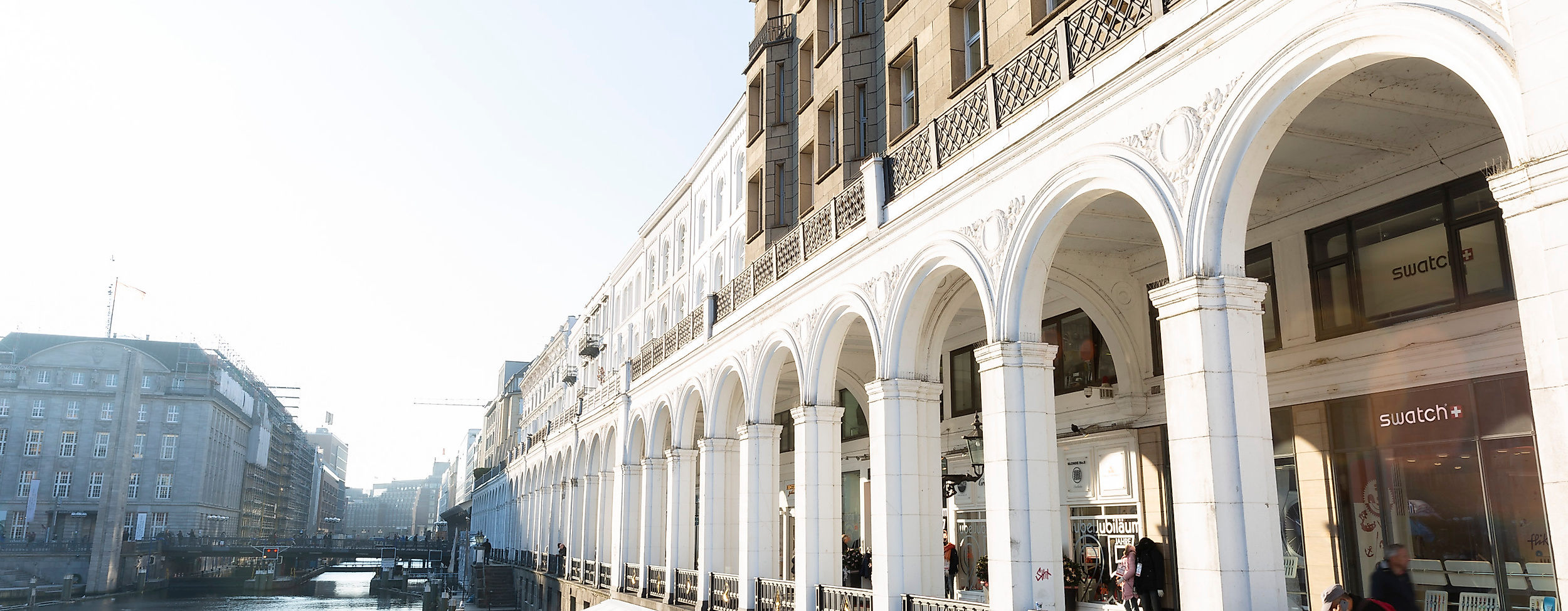 Alsterarkaden in Hamburg mit weißen Säulengängen, Geschäften und Blick auf das Wasser bei strahlendem Sonnenschein