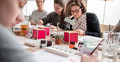 Mehrere Personen arbeiten konzentriert an einem langen Tisch mit Lettering-Vorlagen, Pinseln und Tinte.
