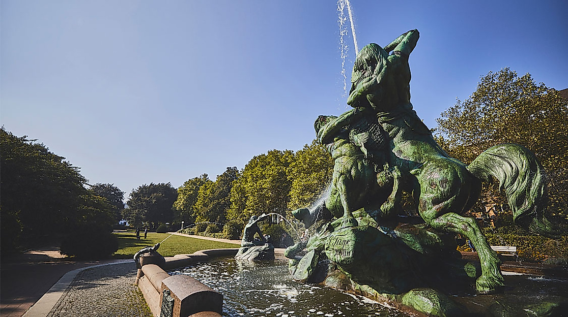 Bronzefigur des Stuhlmannbrunnens mit Wasserfontäne im Altonaer Park, umgeben von Bäumen bei Sonnenschein