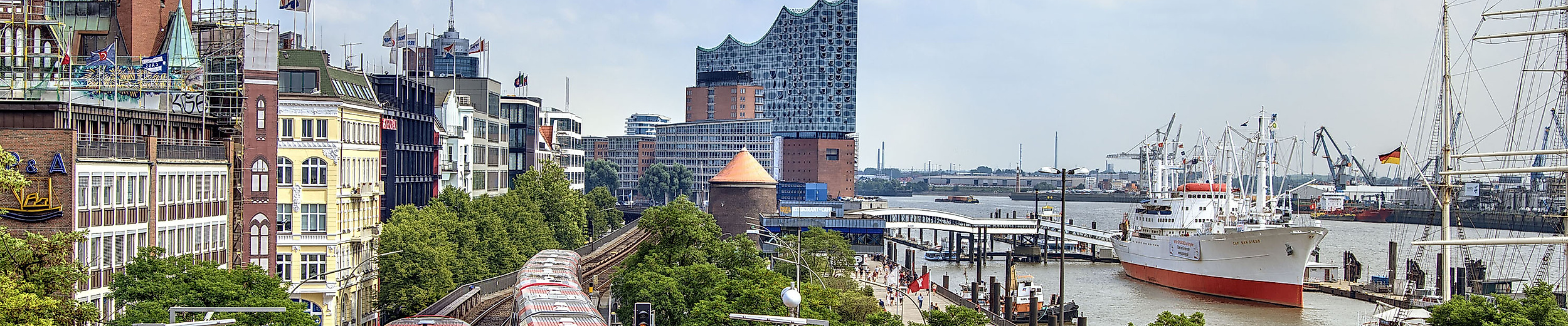 U-Bahn fährt an den St. Pauli Landungsbrücken entlang, im Hintergrund Elbphilharmonie und Museumsschiffe.