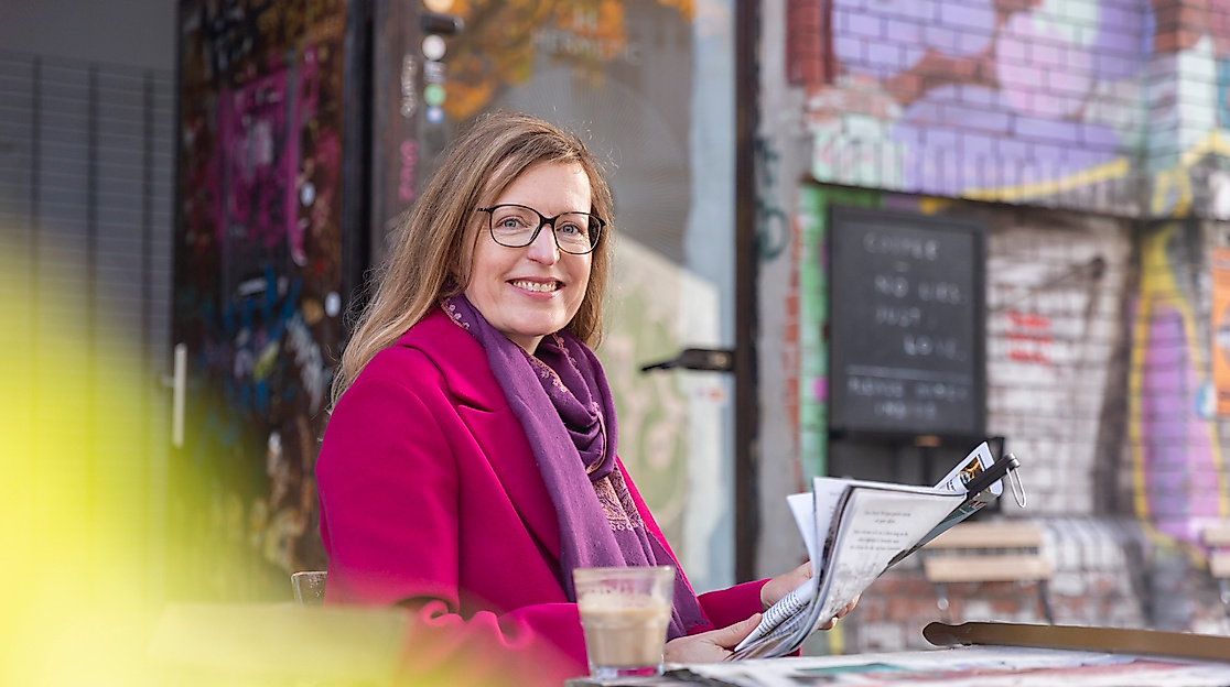 Lächelnde Frau mit Zeitschrift und Getränk im Straßencafé vor bunt besprühter Wand im Hamburger Karoviertel