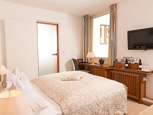Helles Zimmer mit Doppelbett, Holzschreibtisch, Fernseher an der Wand und Fenster mit Vorhängen.