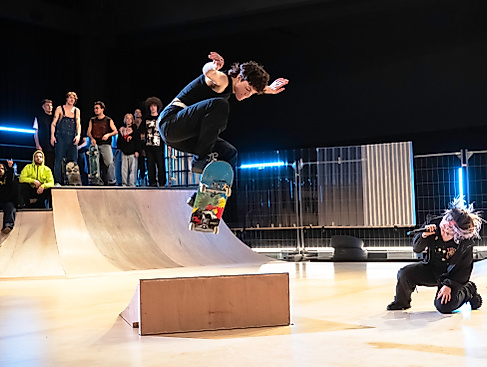A skateboarder dynamically jumps over an obstacle while a performer with a microphone sings in a squatting position and a group of spectators watch in the background.