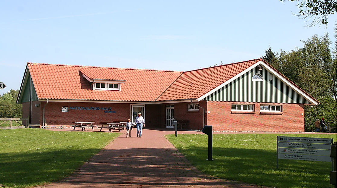 Backsteinbau des Nationalpark-Hauses Neuwerk mit rotem Dach, Besucher:innen und grünem Außengelände