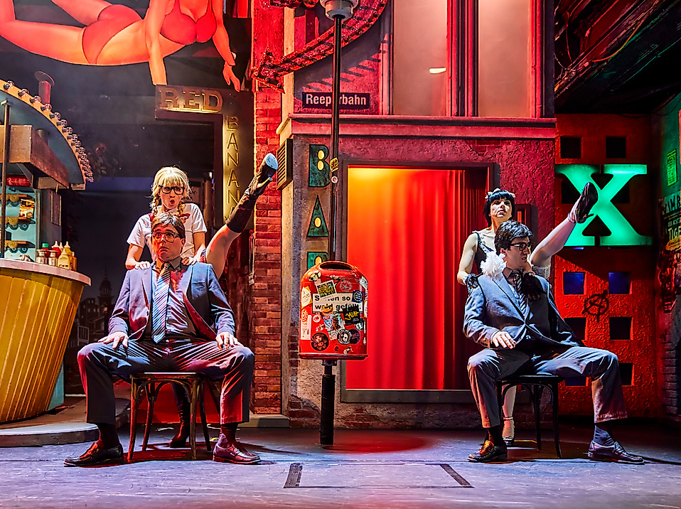 Szenenbild Heiße Ecke: Zwei Männer sitzen auf Stühlen, hinter ihnen steht jeweils eine Frau