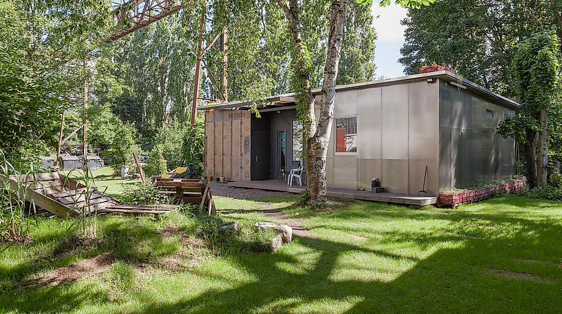 Photo of the Migrantpolitan at Kampnagel. A small one-storey house with a green garden, benches made of wooden pallets, surrounded by many trees.