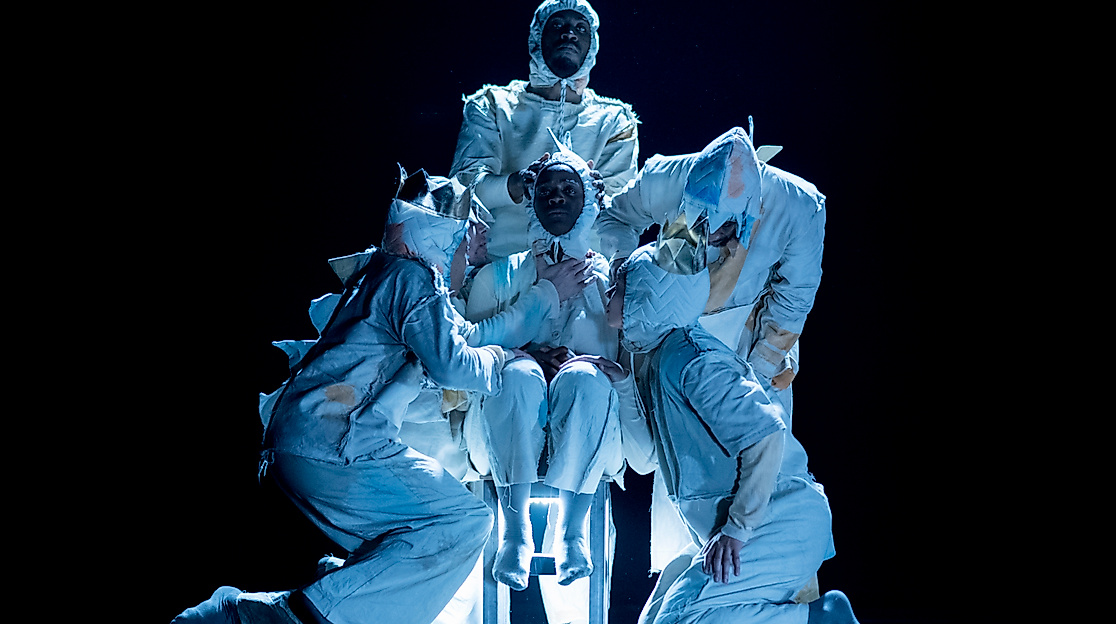 Four performers stand close to a person sitting on a chair. They are all wearing bright costumes with large spikes on the back.