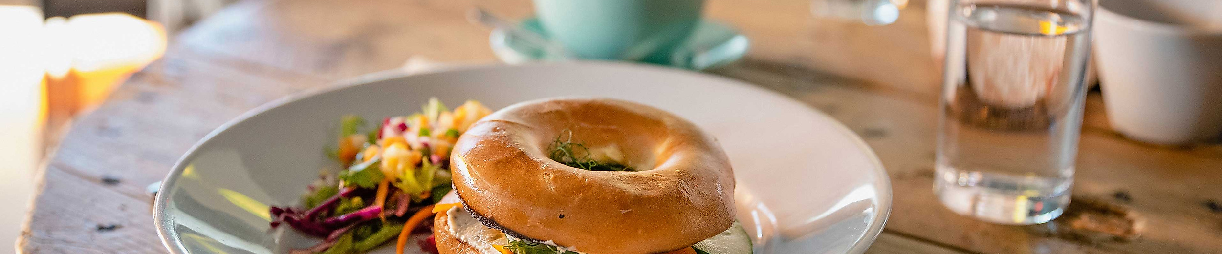 Frühstücksbagel auf weißem Teller, dazu Tasse und Glas auf Holztisch