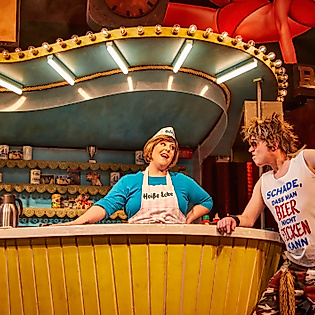 Szenenbild Heiße Ecke: Ein Mann vor dem Tresen redet mit einer Frau hinter dem Tresen