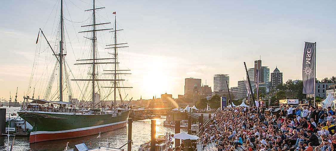 Menschen genießen das Hafenflair an den Landungsbrücken bei Sonnenuntergang – im Vordergrund ein historisches Segelschiff, dahinter das Panorama der Stadt in goldenem Licht.