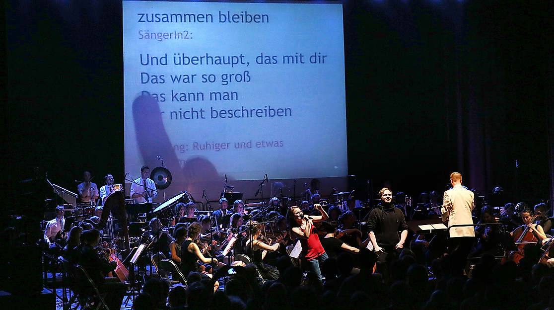 Ein Orchester spielt auf einer Bühne, im Hintergrund ist eine große Leinwand, auf die ein Songtext projiziert wird. Vor dem Orchester steht eine junge Frau, die fröhlich in ein Mikrofon singt.