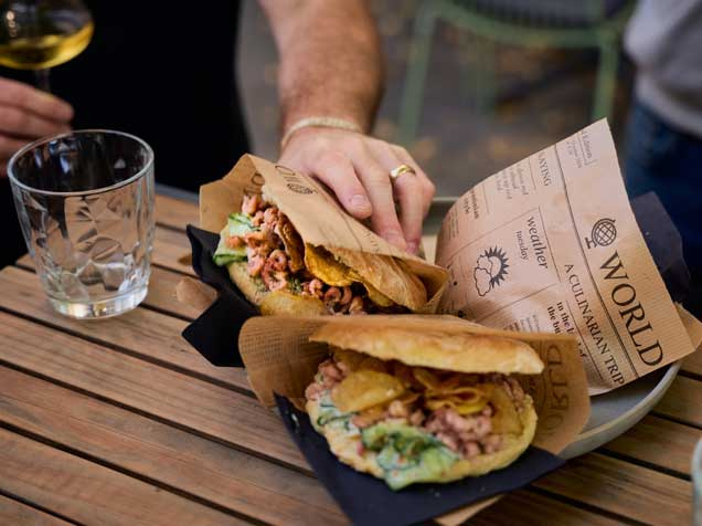 Zwei belegte Pita-Taschen mit frischen Zutaten wie Krabben und Salat liegen auf einem Holzteller, serviert auf einem Holztisch. Eine Person greift nach einem der Snacks. Im Bildhintergrund ein Glas Wasser und ein Glas Weißwein – perfekte Szene für einen genussvollen Stopp bei einem Partnerrestaurant der Hamburg CARD mit Rabatt.