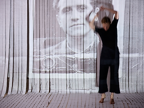 The performer moves around the room with raised arms. Behind them is a curtain with white stripes, on which a man's driver's license photo can be seen.