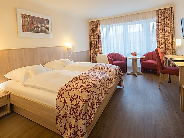 Helles Hotelzimmer mit Doppelbett, zwei roten Sesseln am Fenster und floralen Dekoelementen.