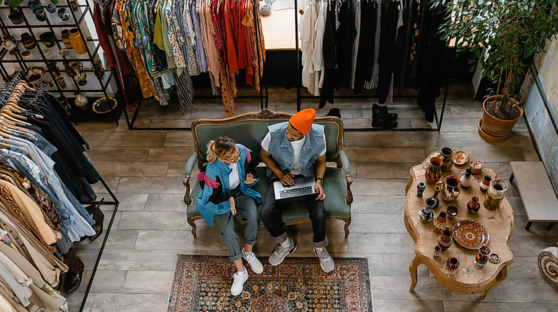 Zwei Personen sitzen entspannt in einem stilvoll eingerichteten Second-Hand-Laden mit vielfältiger Kleidungsauswahl