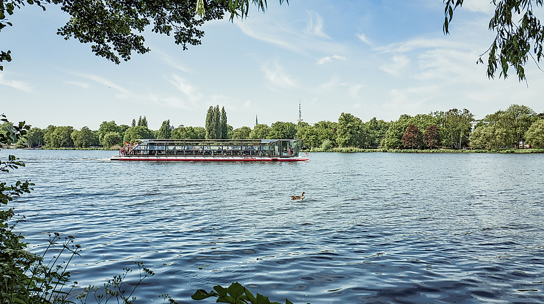 Solarschiff gleitet über die Alster, umgeben von grüner Uferlandschaft und blauem Himmel