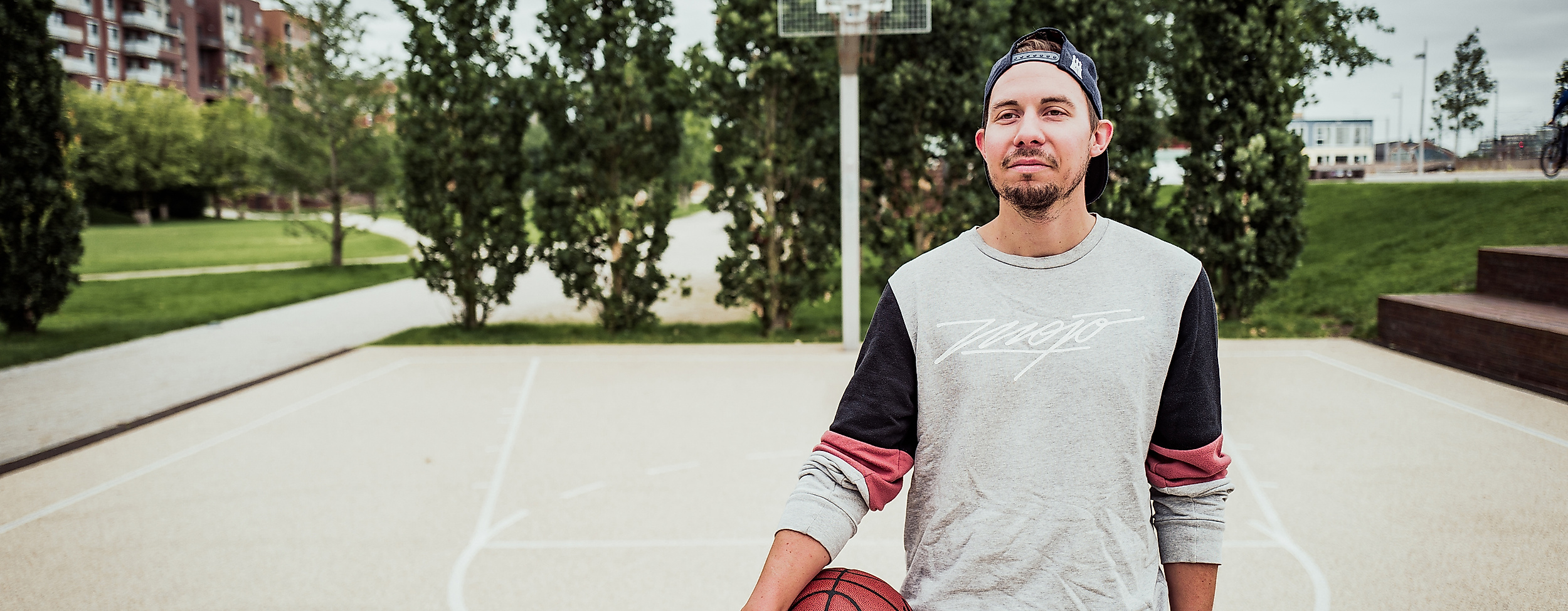 Junger Mann mit Basketball steht auf dem Sportfeld im Lohsepark vor einem Korb und hohen Bäumen