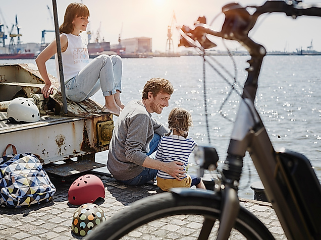 Eine Familie mit Kind sitzt an der Hafenkante, im Vordergrund ist ein Fahrrad zu sehen