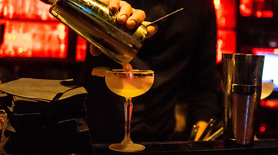 Zwei Cocktails werden an einer Bar bei warmem Licht serviert, daneben Gläser und Barutensilien