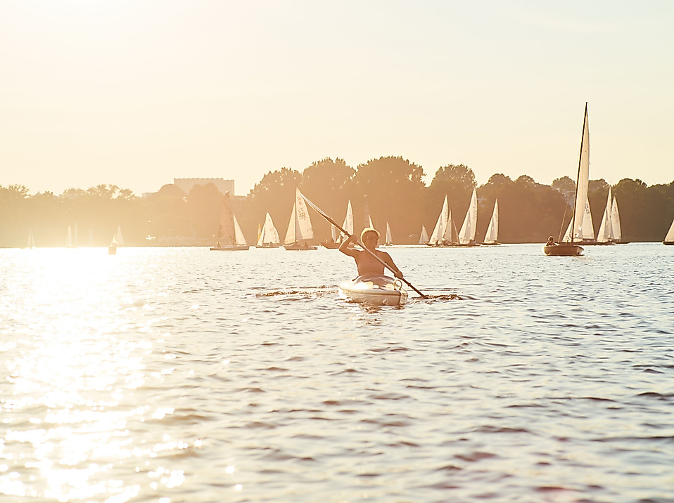 Person paddelt im Kanu auf der Alster bei Sonnenuntergang, umgeben von zahlreichen Segelbooten