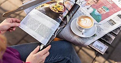 Person liest Zeitung im Straßencafé, auf dem Tisch liegen Cappuccino und offene Zeitungsseiten bei Sonnenlicht