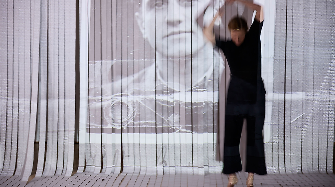 Die performende Person bewegt sich mit erhobenen Armen im Raum. Dahinter ist ein Vorhang aus weißen Streifen, auf den das Führerscheinbild eines Mannes zu sehen ist.