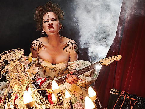 Emma Frankland, eine weiblich gelesene Person mit braunen Haaren steht mit einer E-Gitarre und rockigem Gesichtsausdruck vor einem Vorhang. Im Hintergrund ist Rauch zu sehen, während im Vordergrund ein Kronleuchter ist.