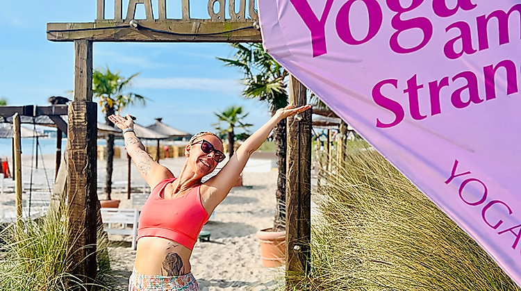 Frau steht am Strand vor den HAFFdüün bei Sonne und mit einem Schild Strandyoga YOGACOAST in Haffkrug an der Ostsee